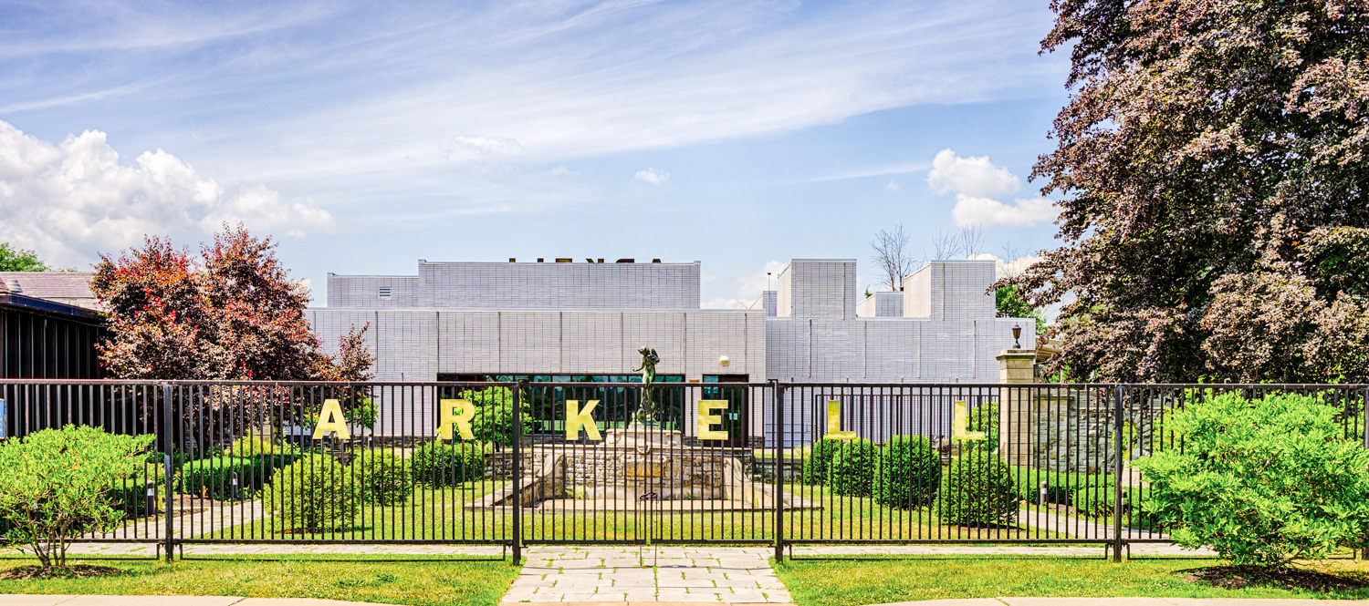 Architectural Photography of the Arkell Museum building with white facade, black fence labeled "ARKELL," sculpture, trees, cloudy sky.